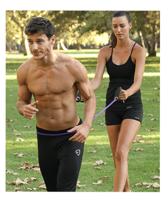 A muscular, shirtless man runs in a grassy park while a woman behind him holds a purple resistance band attached to his waist, providing resistance.