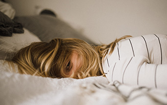 A person with blonde hair lies face down on a white bed, their head mostly buried, with one tired eye visible.